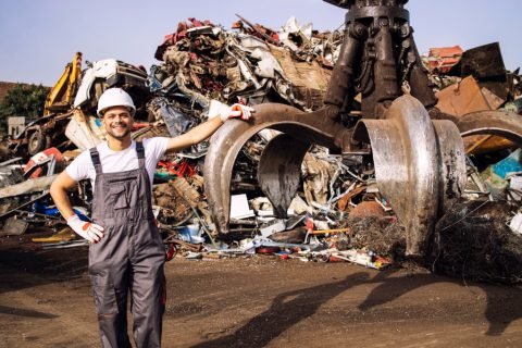 Portrait of worker standing by hydraulic industrial machine with claw attachment used for lifting scrap metal parts in junk yard. Scrap Processing Services in Columbia, SC