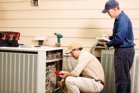 Air conditioner repairmen work on home unit. Blue collar workers. Caucasian and Latin descent repairmen work on a home's air conditioner unit outdoors. They are checking the temperature and taking notes with clipboard.