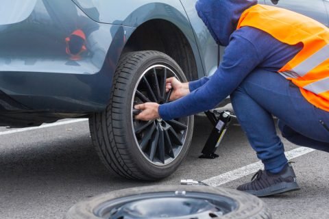 A man in a orange safety vest changes a flat tire on a road. Close-up mans hands to the wheel of a broken car. Replacement of a wheel using skrewdriver, Prague, March, 2020 Tire Change Services In Stockton, CA